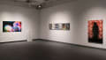 A corner wall inside SVA’s Gramercy Gallery. On the very far left wall is a colorful photograph, and on the right wall is a series of six photographs lined neatly up, and another large image.
