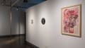 Three pastel drawings of various sizes and colors hang on a wall in the hallway of the gallery. A sculpture in a long strand hangs from the ceiling ahead.