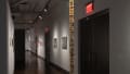 A hallway in SVA Chelsea Gallery, with artwork and sculptural pieces hanging on the left and right walls.