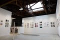Gallery interior with colorful comic and illustration artworks on white walls, a standing monitor, and a wooden beam ceiling with a skylight above.