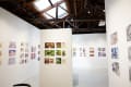 Gallery interior featuring colorful comic and illustration artworks on white walls under a wooden ceiling with exposed beams and a skylight.