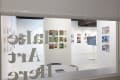 View of a bright gallery interior seen through a window with the words “Make Art Here” in reverse on the glass. The exhibition features colorful illustration and comic-style artworks arranged in neat grids on white walls. A digital display monitor stands on a white pedestal at the center, and a round table with a brochure is visible in the foreground. 