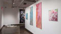 In the hallway before gallery four, two medium sized canvases depicting flowers hang on either side of two long tapestries depicting tall buildings. In the back corner, white plastic octagons are placed around a chair on the floor.