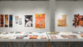 View of the display table in the center of the back gallery and the left hand wall behind it. There are more prints on the wall visible, including one of a quaint tea shop, and a fractured puzzle of a girl surrounded by red and black flowers. 