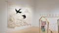 Another view of the west and south walls in the Flatiron Gallery, showing a wooden house-like structure with rope walls on the floor in the foreground. Papers from a book are pinned to the wall on the right, and a large white tapestry with black swallows hangs on the south wall.