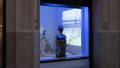 View from outside the window of the Flatiron Project Space. A mixed media sculpture of a bust is in the foreground, directly behind the window. A projection and more colorful sculptures are visible inside. 