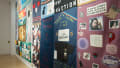 An angled view of the east wall of the SVA Flatiron Gallery, showing two large memorial quilts hanging on the wall. The one to the right has a grid of 8 rectangular memorial patches, and the one to the left has eight memorials of mostly horizontal orientation surrounding two center patches.