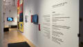 Wall vinyl in the hallway reads “To Love-To Die; To Fight. To Live” along with information about the exhibition. Past the vinyl, there is a monitor and then a large quilt stretching from the top of the wall almost to the floor.