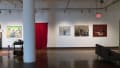 On the left, a room-like installation is set up in the corner of the space, with a large painting, a desk, a candle, and a red velvet curtain. To the right of this installation, three paintings on canvas are hung in a row.