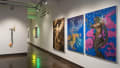 On the right wall of gallery one, three large paintings depict human figures merging with nature in various states. One painting is monotone sepia, while the other two are a vibrant cobalt with pinks and yellows. A mixed media assemblage is visible on the wall to the left.