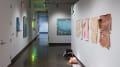 Looking back toward gallery one through the right hallway, some paper works displayed to the immediate right are seemingly peeling off the wall. Sculptures sit on the ground beneath them, while large paintings are visible on the left wall and in gallery 3 beyond them.