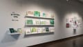 Angled view of the west wall at the SVA Chelsea Gallery. Three shelves of watercolor paintings in blues and greens hang in the foreground, and a small shelf with books and cards hangs below the title, “The Blackthorn Woods, Emma Fisher.”