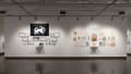 A view of the east wall at the SVA Chelsea Gallery. On the left is a black and white project hung salon style. There is a large black flag with a cat with a sword in white at the top, under the words, “Bonny sets sail.” Another project hung salon-style is on the right, with many colorful framed prints under the title, “For All We Know.”