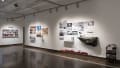 An angled view of the south wall in Gallery 2 shows a project with a large imitation rock on the wall below the title “Crevice.” There are many largely black and white prints hung above it. The the left theres a pin board with postit notes and small cards, and many colorful prints next to it.