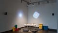 In a corner of gallery one, three objects (a cube, a sphere, and a pyramid) are laid out on a small table. The table is surrounded by floor pillows, and colorful projections glow on both walls.