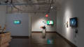 In gallery four, a TV monitor hangs on the wall straight ahead, playing an animated video. Next to it, prints are pinned on the wall, and a pedestal holds a small sculpture. To the left, a large wooden sculpture on the floor is visible in the gallery’s alcove, and to the right, another monitor hangs beside a small shelf.