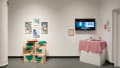 Two works in the exhibition. On the left, four artworks hang above a series of crates. A chessboard balances on top of the crates. On the right, a video plays on a television . Several works of embroidery hang on the walls beside the video, and a plinth sits below with a tank on top of it.