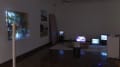 In a dark corner of an art gallery, glowing tv and computer monitors sit on the floor beside two large photographs on the wall. To the left, a projection shines onto a piece with many layers.