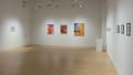 Three brightly colored paintings are centered in the far wall of the gallery. To their right, several small prints are hung in a row. To their left, a framed print hangs alone on a wall.