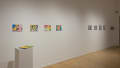 Several colorful prints are hung in a row on the left side of a wall. A book sits on a pedestal in front of them. Several small black and white prints hang on the same wall towards the right.