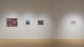 In the gallery, four works hang in a row on a wall. The largest piece is a painting of a checkout counter at a deli. Another smaller piece shows a woman dressed as a geisha looking in a bathroom mirror.