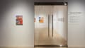 Next to the door of the SVA Flatiron Gallery, wall vinyl displays the exhibition title “Around and Again” along with the artists’ names. One painting hangs to the left of the glass door, and more artworks are visible inside.