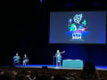 A man gestures to a projection while giving a talk on a stage