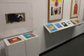Two children’s books are present on a pedestal. To their right, a series of small collages sit on a shelf beneath two large framed works.