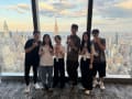 Six people pose in front of a large window with a stunning view of the NYC afternoon skyline.