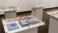 Four white pedestals, each containing multiple printed books, and covered in large sheets of clear plexiglass.
