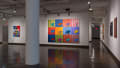 Two paintings hang from a large wall in center of gallery space with hallways on either side, the rightmost of which contains several paintings.