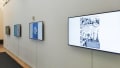 Installation view of artwork on four monitors in a white hallway, the closest of which shows a black and white comic with the text "Satire On Fair Ladies" beside it.