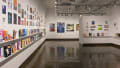 Multiple views of SVA Chelsea Gallery. There is illustrations and paintings hung on the walls. There are multiple shelves throughout the gallery with books places on them.