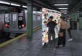  A 3D rendered image of an NYC subway interior is shown. A subway platform is visible, with a train stopped at the station on the left side of the image.On the platform to the right, a large panel is shown with handwritten text at the top of the panel. The panel shows a map of the NYC subway with images of people’s faces pasted on different parts of the map