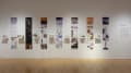 Installation image showing a straight view of a gallery wall with seven building models on shelves, and information banners for each to the right.