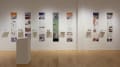 Installation image showing a straight view of a gallery wall with seven building models on shelves, and information banners for each to the right. There is a pedestal on the floor with a book on top.