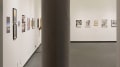Entryway to gallery space, bisected by a support column. Many artworks hang across two walls. 