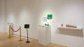 Against one wall of the SVA Flatiron Gallery, a shelf holds several small sculptures of an anatomical heart. To the left of the shelf, colorful paper works sit on and above a pedestal. Further left, a stanchion with a red velvet rope protects a row of paper stars that lay on the ground.