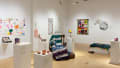 A wider view of the gallery shows a chair with a knit blanket across it, surrounded by pedestals that hold small sculptures. One wall holds colorful drawings and a mirror with a vibrant frame.