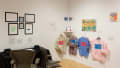 In one corner of the gallery, there are two office chairs facing a table and several framed works hanging above it. To the right, a row of sweatshirts are printed with pink and blue text. Around them hang various drawings and small sculptures.