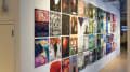 Four rows of colorful, illustrated posters are displayed in four rows on a white gallery wall. The posters are each about 11 x 14 inches.