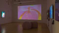 View inside gallery space showing one large projection on the back wall and monitors playing colorful abstract videos on the left and right walls.