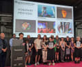 A person speaks at a modium with award recipients gathered around and a projection screen showing the awarded project behind them