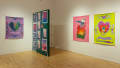 Installation image featuring a painted wooden wall erected on the floor with prints attached, and 3 paintings on opposing walls in purple, green and pink.