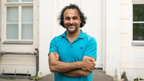 Image of Kader Attia, a man standing in front of a white door with his arms crossed