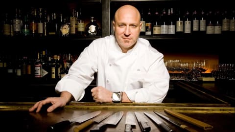 Portrait of Tom Colicchio behind a bar with knives on the counter, all pointed towards him.