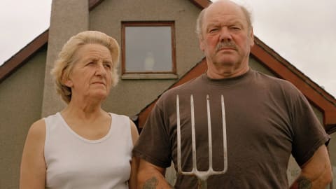 An older man and woman stand in front of their farmhouse on a gray day. The man is holding a pitchfork prongs up and staring at the camera. The woman is staring off in the distance.
