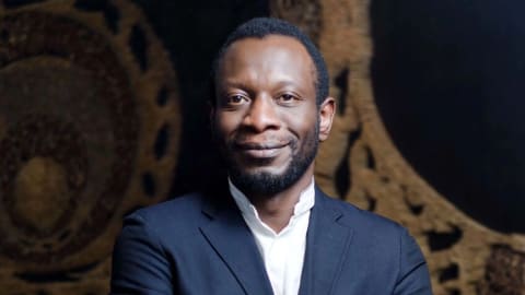 Color portrait of Azu Nwagbogu, wearing a white shirt, navy jacket and jeans. He's smiling and his arms are crossed. 