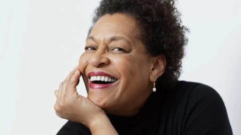 A black woman in a black turtleneck smiling in front of a white wall, resting her face in her hand