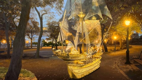 Oil painting of a ghostly ship in a modern-day park in early Autumn. The ship is a light yellow/white with large translucent sails.